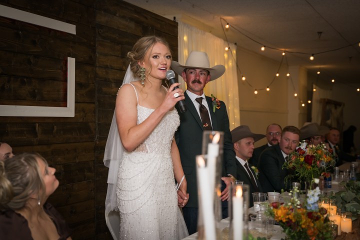 Bride in white dress speaks into microphone at wedding reception, groom in cowboy hat beside her.
