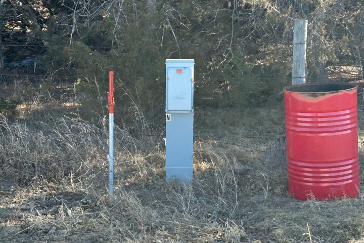 Metal pole with box and red barrel in grassy area near trees.